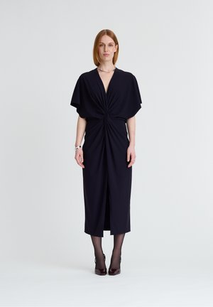 Frau mit glatten roten Haaren, die ein schwarzes Midikleid mit verdrehtem Vorderteil, schwarze Strumpfhosen und dunkle High Heels trägt und vor einem einfarbigen Hintergrund steht.