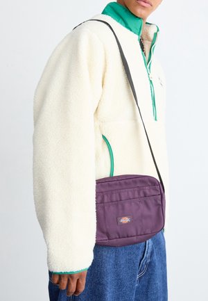 Person trägt eine weiße Fleecejacke mit grüner Einfassung, blaue Jeans und eine kleine lila Umhängetasche mit einem Logopatch.