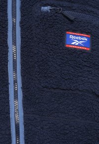 Tmavě modrá fleecová bunda s plným zipem a šedými akcenty. Obsahuje zipovou náprsní kapsu a nášivku loga Reebok. Měkká textura.