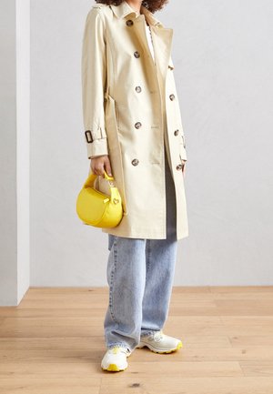 Persoon draagt een beige dubbelgeknoopte trenchcoat, lichtblauwe spijkerbroek, witte sneakers met gele accenten, houdt een kleine felgele handtas vast.