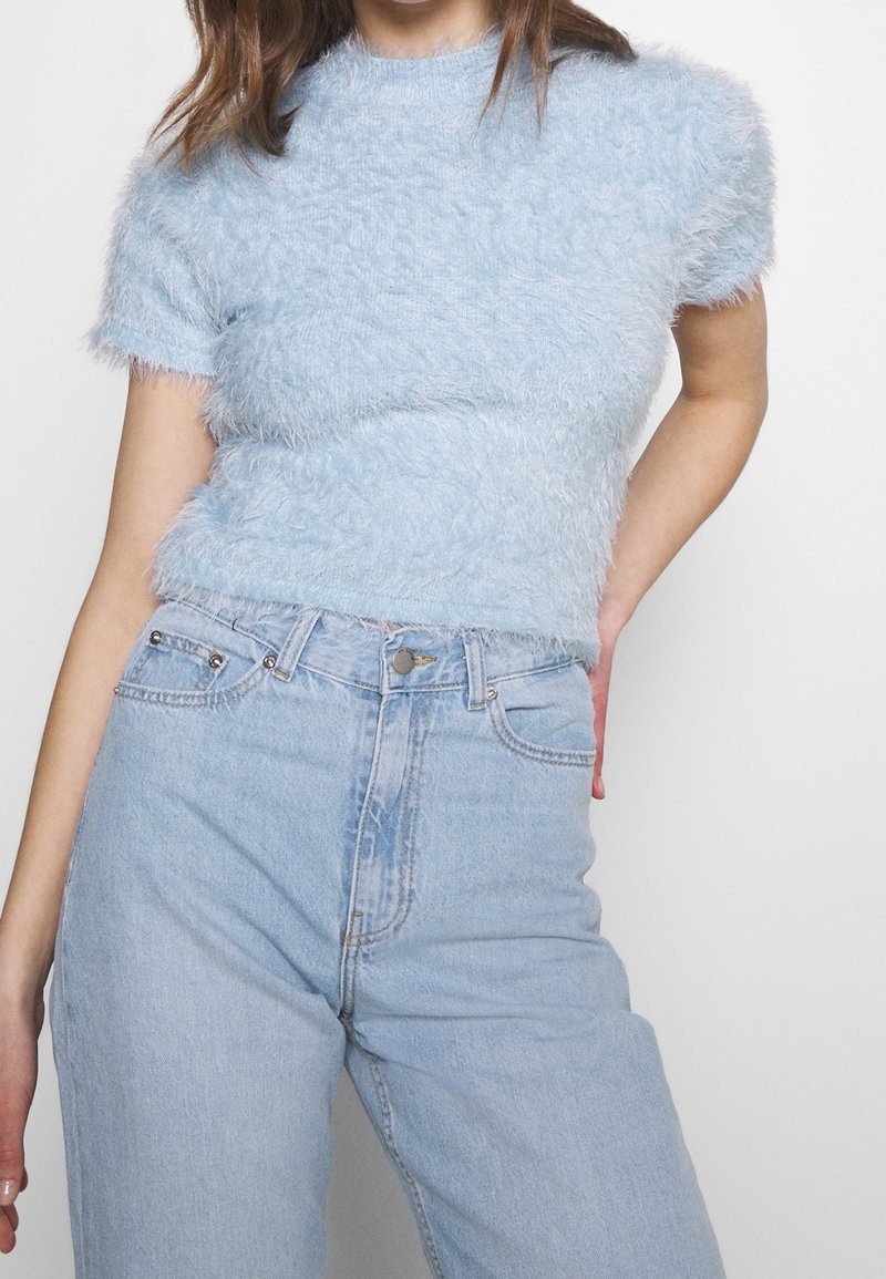 Femme portant un haut bleu clair pelucheux à manches courtes associé à un jean en denim bleu clair taille haute, debout avec une main sur la hanche.