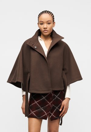 Young woman with braided hair wearing a brown cape jacket over a white top and a dark plaid skirt with buttons.