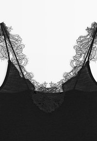 Camisole nero con delicato bordo in pizzo floreale lungo la scollatura e sottili doppie spalline su sfondo bianco.