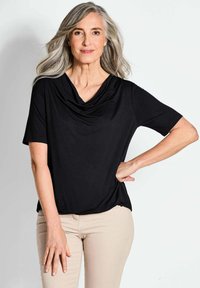 Schwarzes Top mit Kapuze und kurzen Ärmeln, aus weichem Stoff gefertigt. Verfügt über eine lockere Passform und ein drapiertes Design, kombiniert mit hellbeigen Hosen.