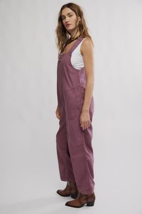 Lila Baumwoll-Latzhose mit bequemer Passform, großer Fronttasche und verstellbaren Trägern. Kombiniert mit braunen Stiefeletten und einem weißen Tanktop.