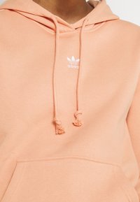 Světle broskvová mikina s kapucí, klokáním kapsou, šňůrkami s třásněmi a bílým logem Adidas na hrudi. Hladká textura, ležérní design.