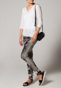 Weißes, knopfbares Oberteil mit 3/4-Ärmeln; gemusterte graue und schwarze Hose mit glänzender Oberfläche; schwarze Umhängetasche; schwarze Sandalen.