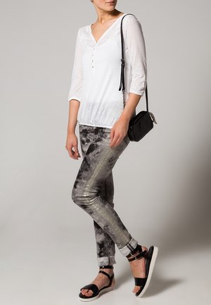 Haut blanc à boutons avec des manches 3/4 ; pantalons gris et noirs à motifs avec une finition brillante ; sac bandoulière noir ; sandales noires.