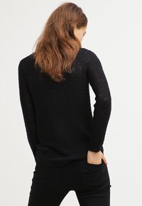 Maglione nero lavorato a maglia con maniche lunghe, tessuto testurizzato, scollatura rotonda e orlo a coste. Il modello ha le mani nelle tasche, visto da dietro.