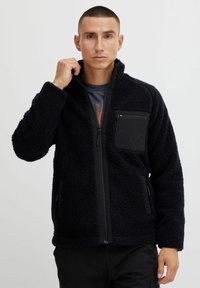 Vezelige zwarte fleece jas met hoge kraag, ritssluiting aan de voorkant en een borstzak. Heeft zijzakken en een gestructureerd oppervlakte.