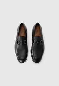 Mocassins en cuir noir avec une texture lisse, bout carré et détail de boucle en métal décoratif sur le devant. L'intérieur présente une semelle intérieure subtilement étiquetée.
