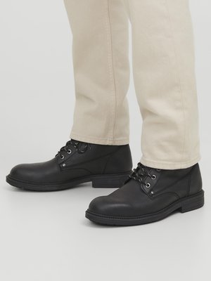 Botas de ante negras hasta el tobillo con punta redonda, ojales de metal y superficie texturizada, combinadas con pantalones de color claro.