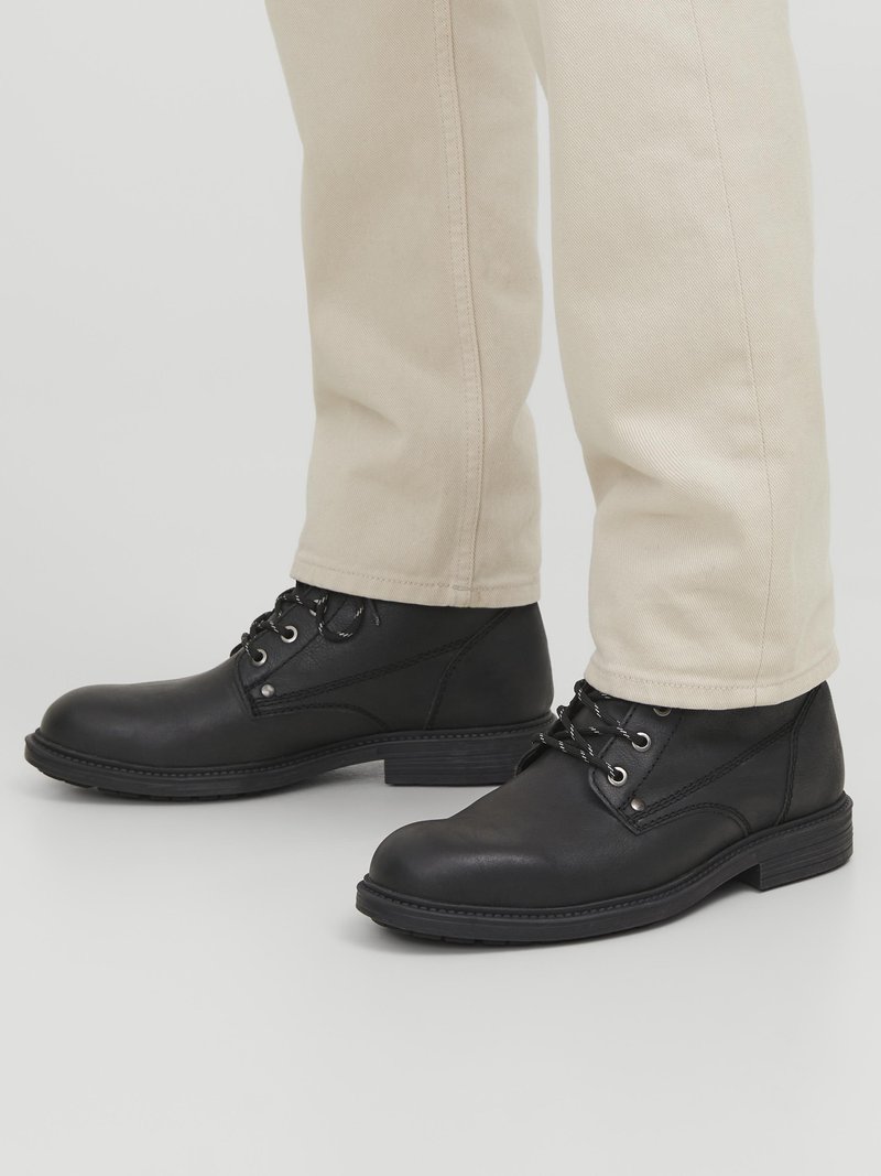 Botas de ante negras hasta el tobillo con punta redonda, ojales de metal y superficie texturizada, combinadas con pantalones de color claro.