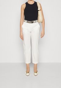 Czarna prążkowana koszulka na ramiączkach, białe dopasowane spodnie, beżowy pasek z klamrą, kremowe czółenka z ostrym noskiem i złotymi akcentami oraz beżowa torba na ramię.