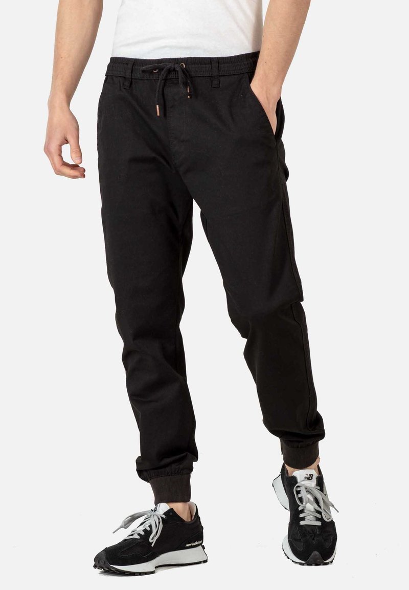 Zwarte joggingbroek met een elastische tailleband, trekkoord, zijzakken, geribbelde boorden en een relaxte fit; gecombineerd met zwart-witte sneakers.