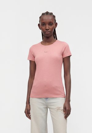 Kvinde med fletninger iført en tætsiddende lyserød T-shirt og lyse, revnede jeans, stående foran en ensfarvet, lys grå baggrund.
