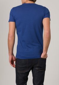 Blå bomullströja med rund halsringning och korta ärmar, sedd bakifrån, i kombination med mörka denimjeans. Ren och enkel design.