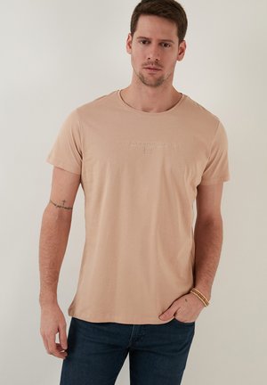 Helle beige Baumwoll-T-Shirt mit kurzen Ärmeln, Rundhalsausschnitt und geprägtem Textdetail. Kombiniert mit dunkelblauen Jeans.