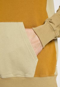 Sweat à capuche bicolore marron clair et moutarde avec une poche avant. Le matériau semble doux, doté de poignets côtelés et d'une étiquette sur le côté.