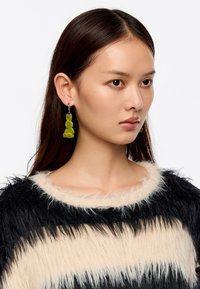 Pendientes de resina verde en forma de gota con un diseño circular y alargado, suspendidos de un aro de plata, llevados por un modelo con cabello largo y oscuro.