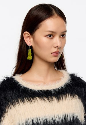 Pendientes de resina verde en forma de gota con un diseño circular y alargado, suspendidos de un aro de plata, llevados por un modelo con cabello largo y oscuro.