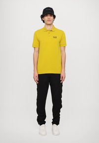Geel polo shirt met drie knopen en EA7-logo, gecombineerd met zwarte sweatpants met zijstroken en witte sneakers. Zwarte pet op het hoofd.