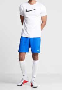 Camiseta deportiva blanca de manga corta con el logo negro de Nike, combinada con pantalones cortos azul brillante, calcetines blancos y zapatillas deportivas blancas con detalles rojos.