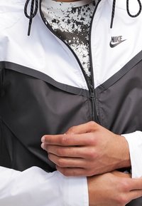 Svart och vit Nike-jacka med dragkedja fram, elastiska ärmar och dragsnodd i huvan, lager på lager över en fläckig svart och vit tröja.