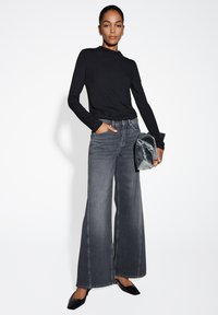 Schwarzer langärmeliger Oberteil, graue weit geschnittene Jeans mit sichtbaren Fronttaschen und schwarze spitze Ballerinas. Transparente Clutch in der Hand.