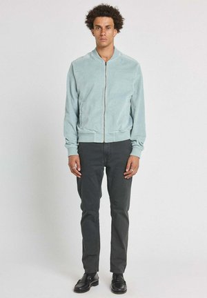 Jeune homme debout, portant une veste zippée bleu clair, un pantalon gris foncé et des mocassins noirs, sur un fond clair uni.