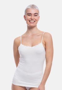 Persona sorridente con capelli platino corti che indossa un top di pizzo bianco su uno sfondo chiaro monotono.