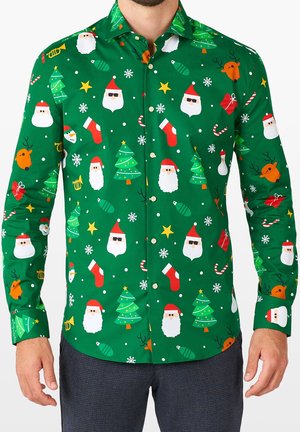 Homme portant une chemise verte à boutons avec un motif sur le thème de Noël, comprenant des visages du Père Noël, des sapins de Noël, des rennes, des cadeaux et des flocons de neige.