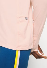 Helles rosa Langarmoberteil mit einer Seitentasche. Glatte Textur, runder Ausschnitt und leicht abgerundeter Saum. Blaue Leggings mit gelben und pinken Akzenten.