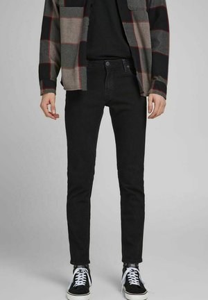 Man wearing black skinny jeans, black shirt, gray and red plaid jacket, and black sneakers with white laces, standing against gray background.