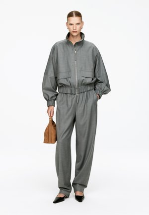 Gray, oversized zip-up jacket with elastic waistband, paired with loose-fitting gray trousers. Accessories include a brown handbag and pointed black shoes.