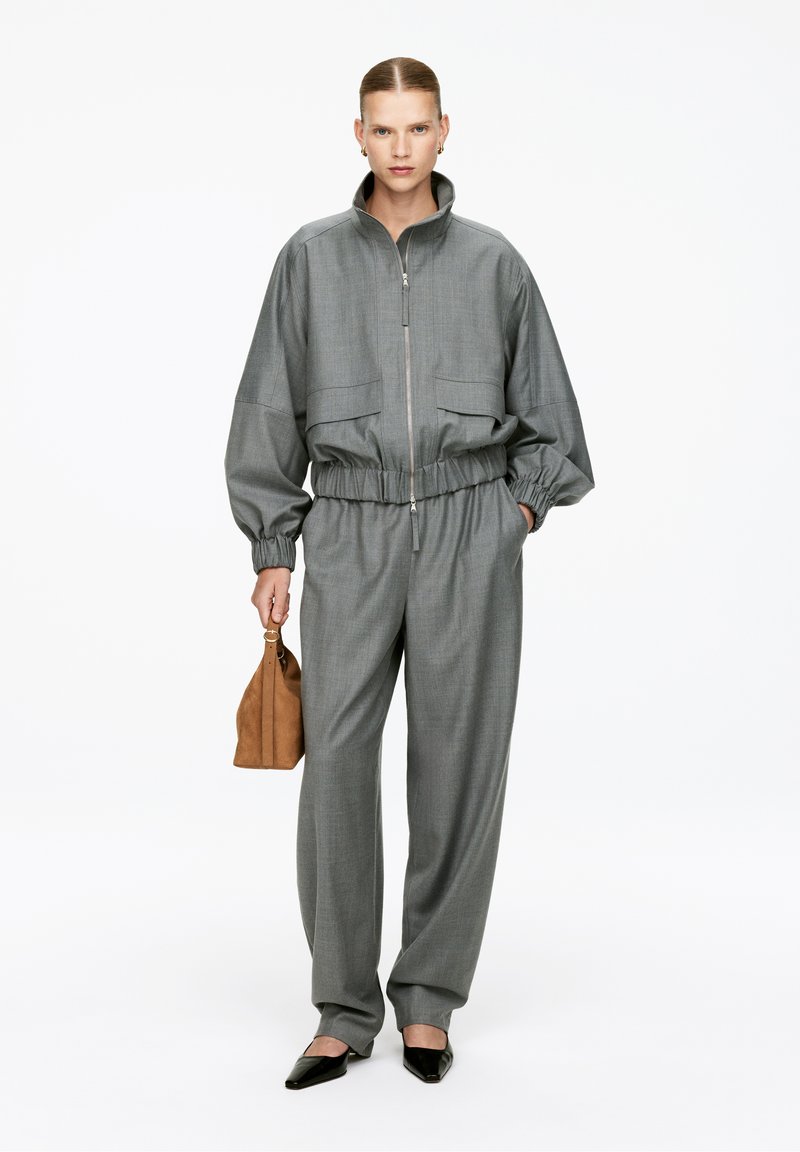 Giacca grigia oversize con zip e cintura elastica, abbinata a pantaloni grigi larghi. Gli accessori includono una borsa marrone e scarpe nere a punta.