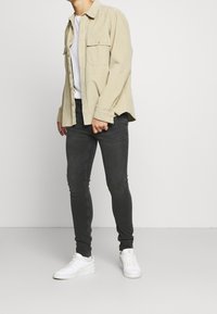 Světle béžová košile na knoflíky s dvěma náprsními kapsami, nošená přes bílý tričko. V kombinaci s tmavými džíny a bílými teniskami. Jednoduchý design.