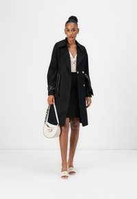 Zwarte trenchcoat met een dubbelbreasted voorkant, grote kraag en brede mouwen. Gecombineerd met een beige top, zwarte rok, beige sandalen en een witte handtas.