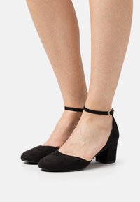 Black suede block-heel shoes with a rounded toe and ankle strap, featuring a small metallic buckle. Smooth texture, minimalistic design.