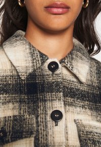Koszula na guziki w kratę w kolorze kremowym i czarnym, wykonana z fakturowanej tkaniny. Posiada kołnierzyk, zapięcia na guziki oraz kieszeń na piersi.