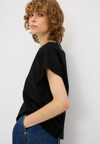 Top corto nero a maniche corte con una vestibilità rilassata, orlo curvo e tessuto testurizzato. Dettaglio della cerniera sul retro di rilievo. Abbinato a jeans in denim blu.