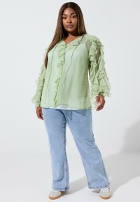 Blusa di chiffon verde chiaro con maniche volants, scollatura a V, texture stratificata, abbinata a jeans a gamba larga di blu chiaro e sneaker bianchi.