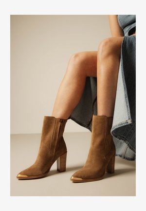 Femme portant des bottines en daim marron à talons blocs et embouts dorés, associées à une jupe en jean.