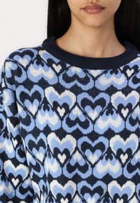 Knit sweater featuring a pattern of hearts in shades of blue, navy, and white, with a ribbed navy crew neckline and textured fabric.