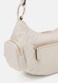 Sac à épaule en nylon beige avec quincaillerie dorée, doté d'un design incurvé, de poches zippées et d'un compartiment latéral arrondi.