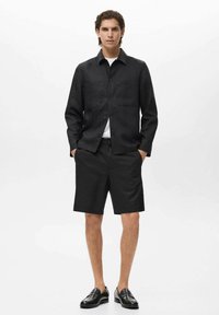 Chemise noire à manches longues avec deux poches avant, assortie à un short noir. Le mannequin porte des mocassins noirs, l'ensemble présente un design minimaliste.