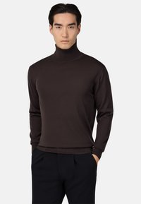Mlad moški s temnimi lasmi, oblečen v prilegač turtleneck pulover v temno rjavi barvi in črne hlače, z rokama v žepih, nevtralen background.