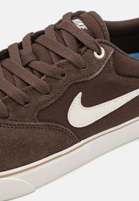 Barna velúr és vászon sneaker fehér Nike swoosh-zal, kerek fűzőkkel és gumitalppal. Texturált felülettel és varrási részletekkel rendelkezik.