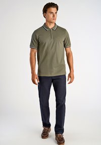 Olivgrön polo skjorta med dragkedja, kontrasterande kant på kragen och ärmarna, tillsammans med mörkblå byxor och bruna mockasiner.