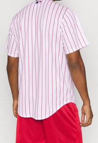 Biała koszula z krótkim rękawem z cienkimi czerwonymi pionowymi paskami. Posiada zaokrąglony dół i małe logo z tyłu na szyi. Połączona z czerwonymi szortami.